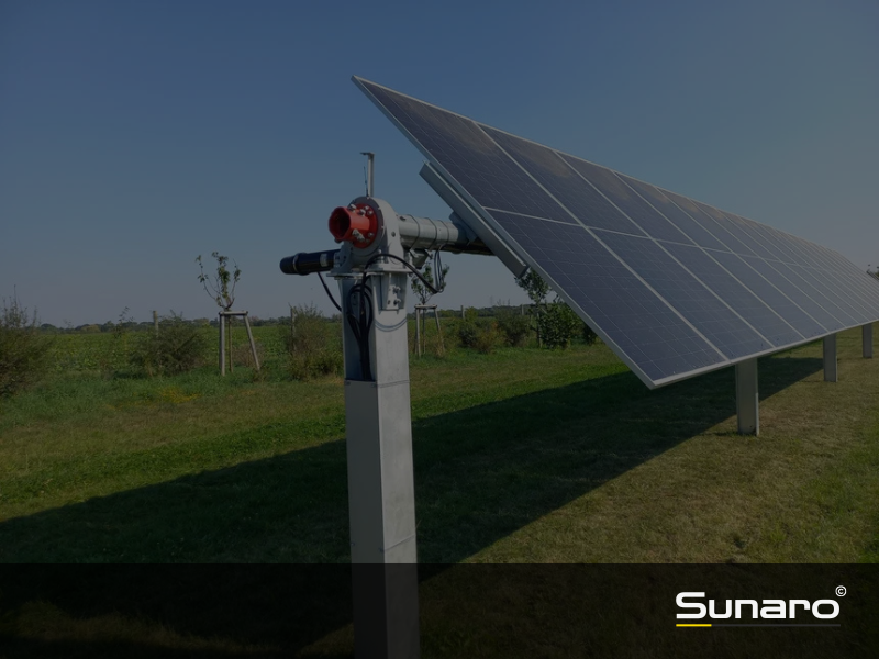Solar-Tracker-Anlage mit einachsiger Nachführung auf einer Wiese, ausgerichtet zur Sonne, im Hintergrund Bäume und offene Landschaft – Sunaro-Logo im Vordergrund
