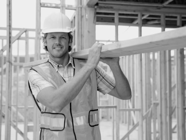 Bauarbeiter mit Schutzhelm trägt Holzbalken auf der Baustelle